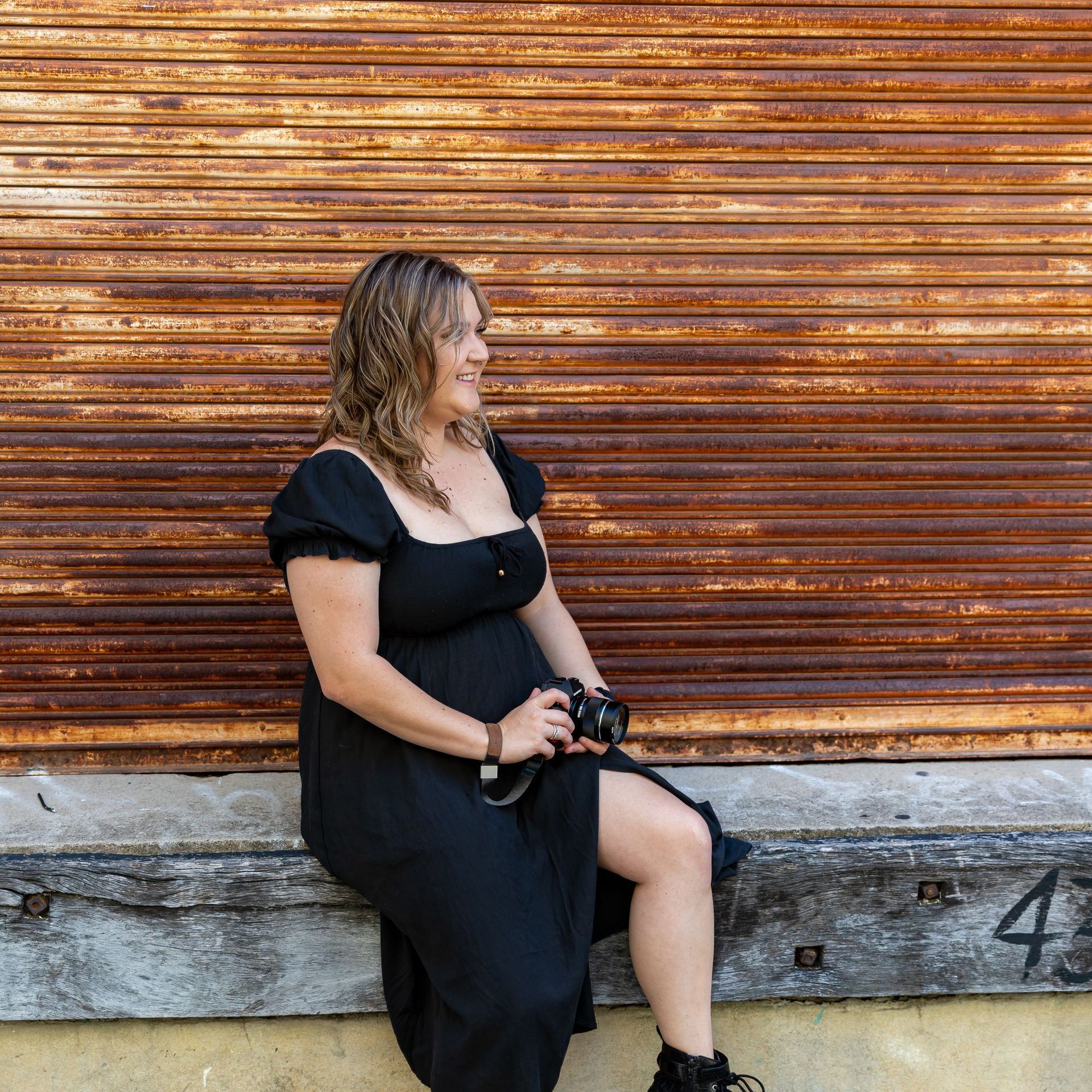 A woman in a black dress is sitting on a bench holding a camera.