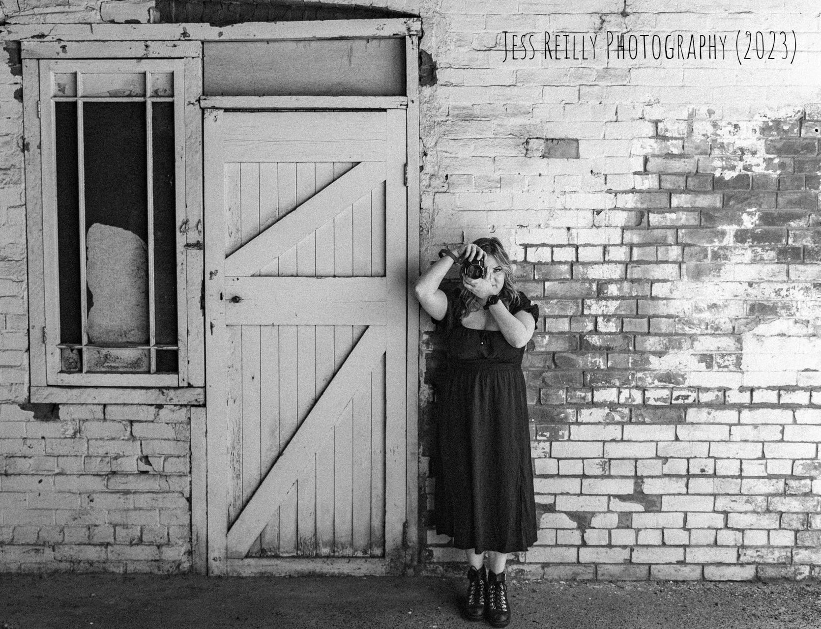 Woman in black dress photographing a brick building.  A window with bars and a wooden door are to the left.