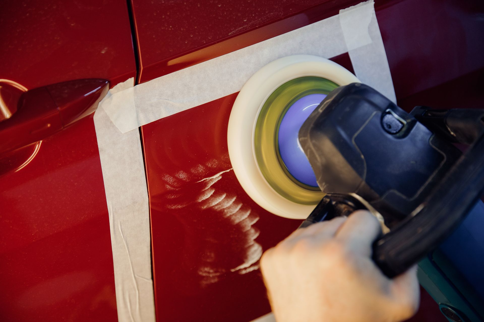 Person using a power polisher on a red car door, taped off for protection.