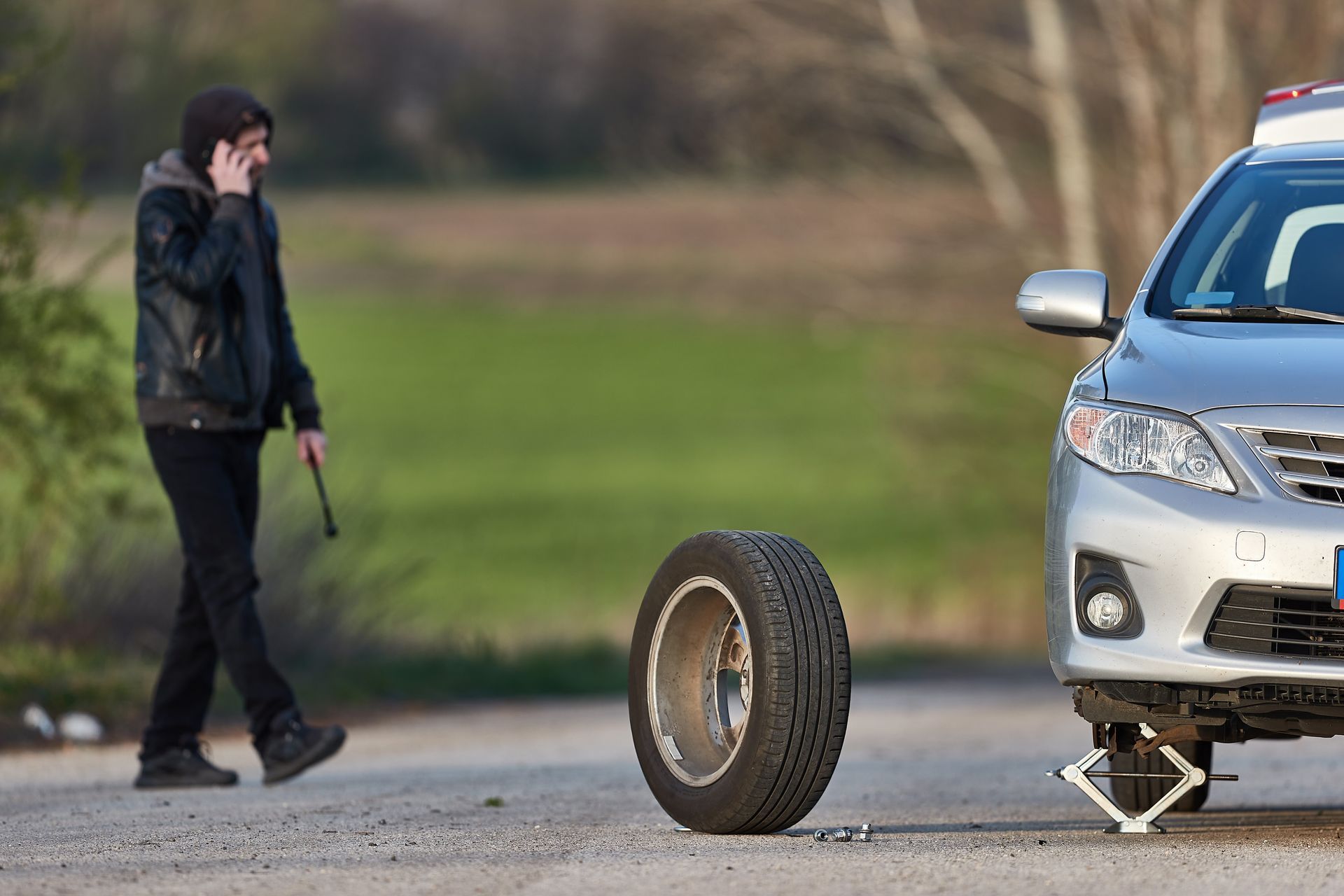 Man on phone beside a car with a flat tire; spare tire on the ground, jack supporting the car.
