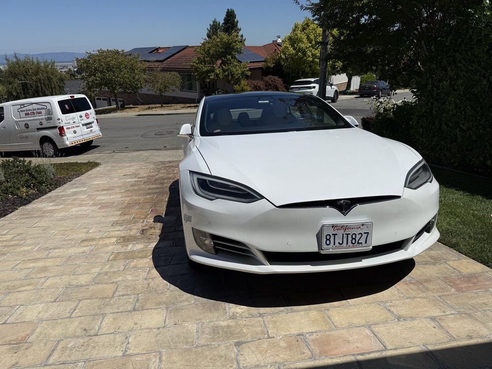White Tesla sedan parked on a brick driveway in front of a house on a sunny day.