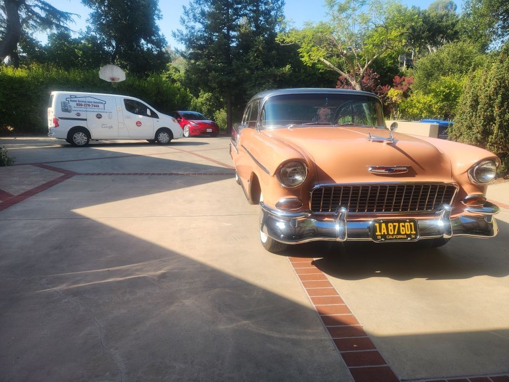 Orange classic car in a driveway, with a service van and red car in the background on a sunny day.