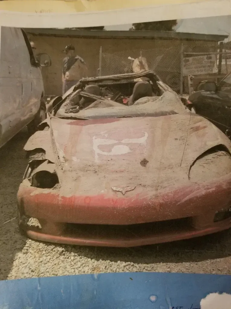 Red convertible car covered in dirt, heavily damaged. Two people stand nearby in an outdoor junkyard setting.