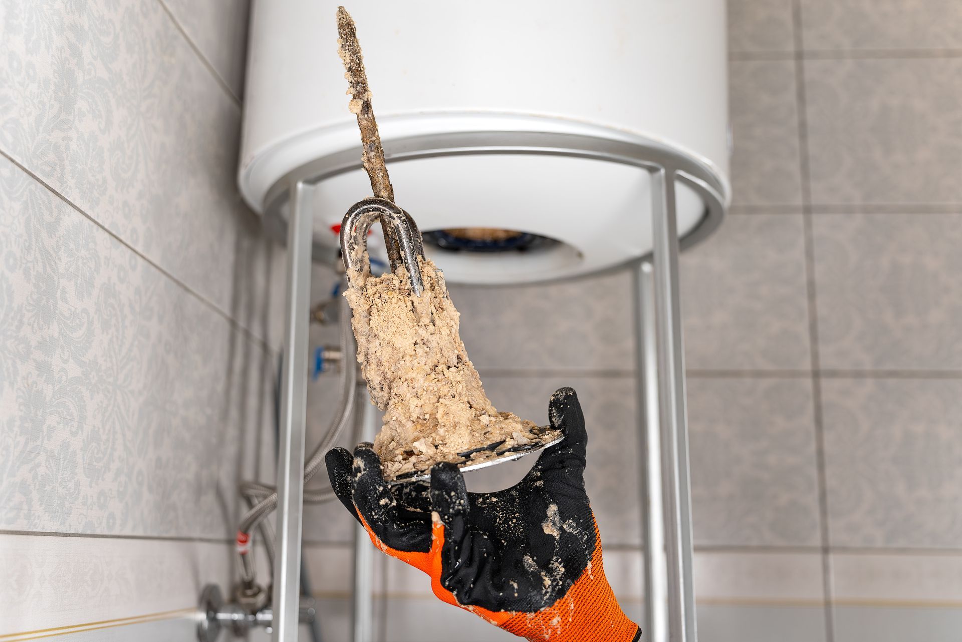 Gloved hand holding a water heater element covered in thick limescale buildup.