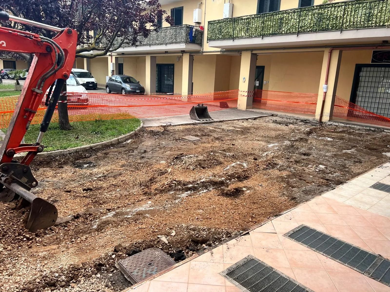 Un escavatore rosso è fermo accanto a un cantiere sterrato di fronte a un condominio a più piani.