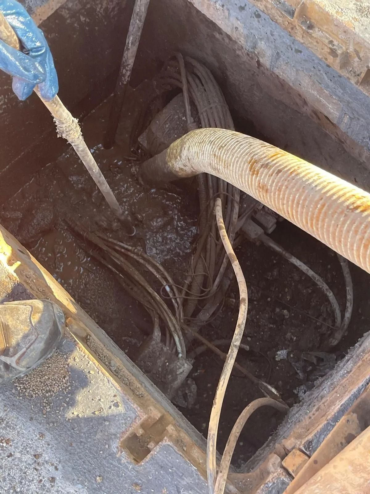 Una mano guantata utilizza un attrezzo per rimuovere il fango da un pozzetto di servizio sotterraneo contenente diversi cavi e un tubo di aspirazione.