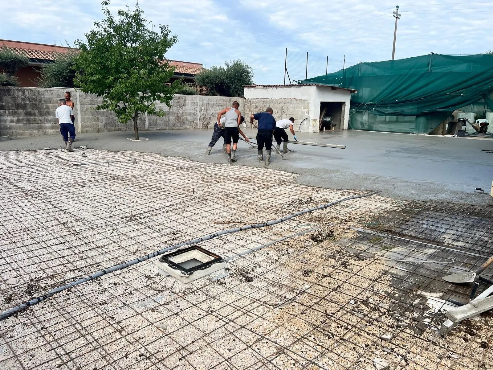 Gli operai edili stendono cemento fresco su un'area di preparazione del cortile, caratterizzata da una griglia metallica su una base di ghiaia.