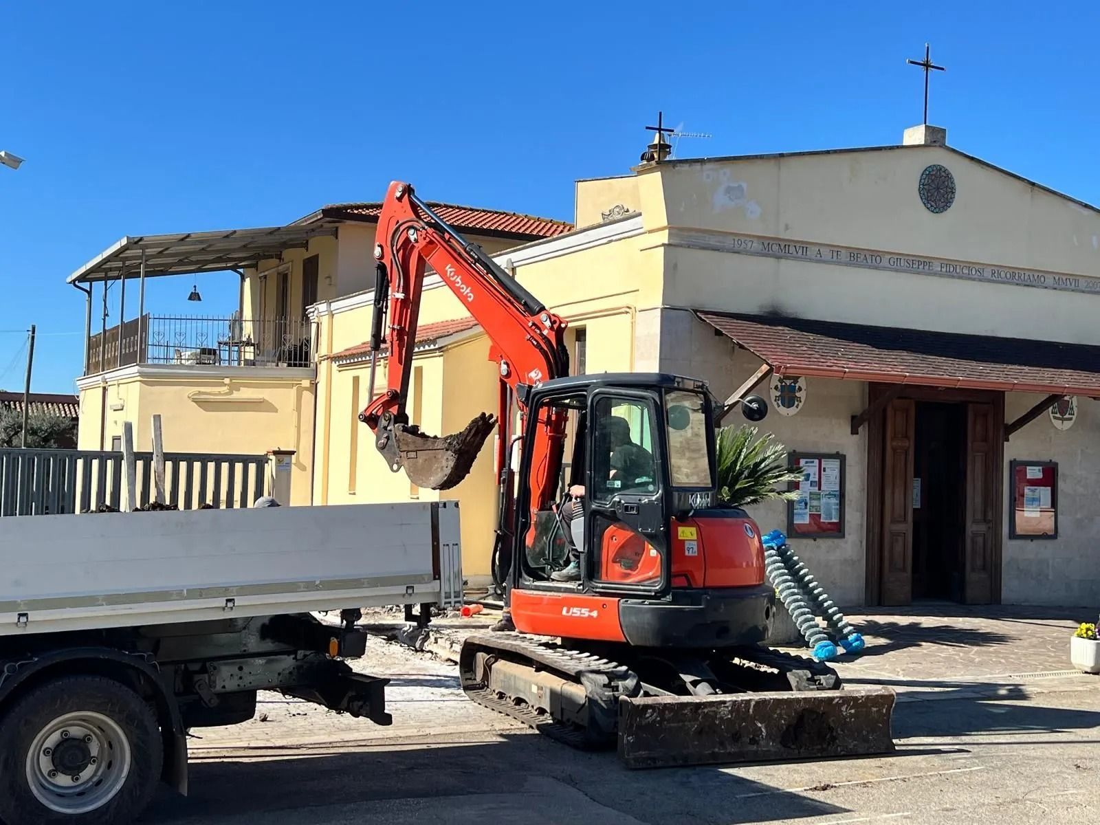 Un escavatore arancione parcheggiato davanti a un edificio in pietra chiara con una croce sul tetto, sotto un cielo azzurro e limpido.