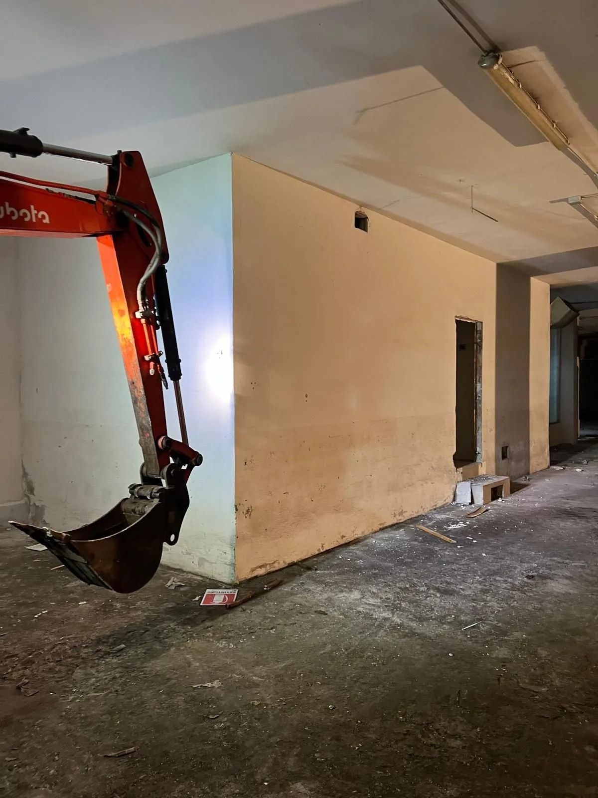 Un braccio di escavatore Kubota arancione posizionato all'interno di un cantiere edile coperto, poco illuminato e ancora incompiuto, con una parete chiara.