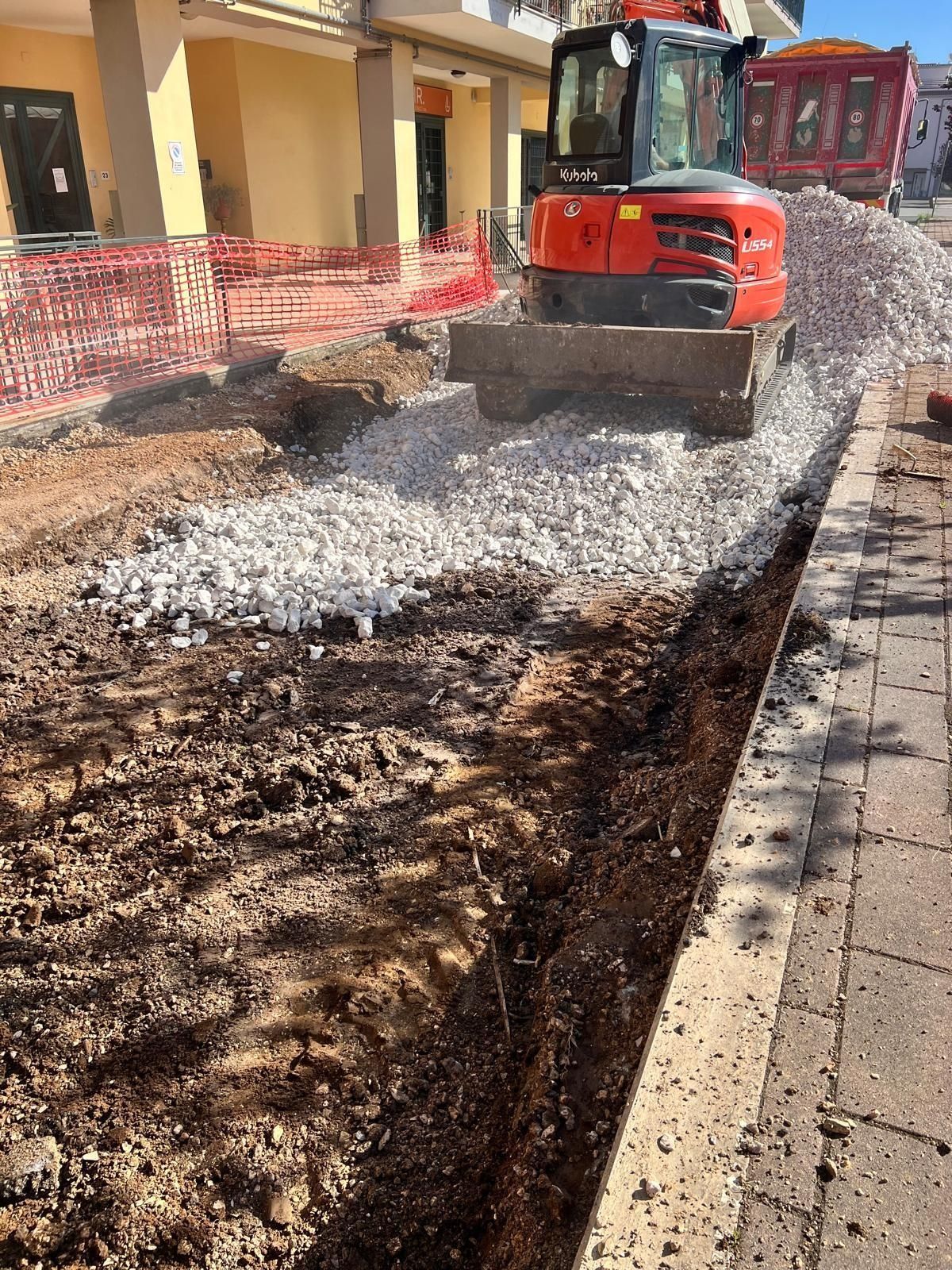 Un escavatore rosso è al lavoro in un cantiere, livellando un cumulo di ghiaia bianca accanto a un marciapiede e a un edificio.