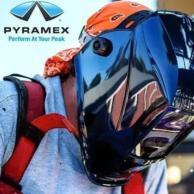 Hombre que lleva un casco de soldador negro, un pañuelo naranja y un arnés de seguridad, con el logotipo de Pyramax.