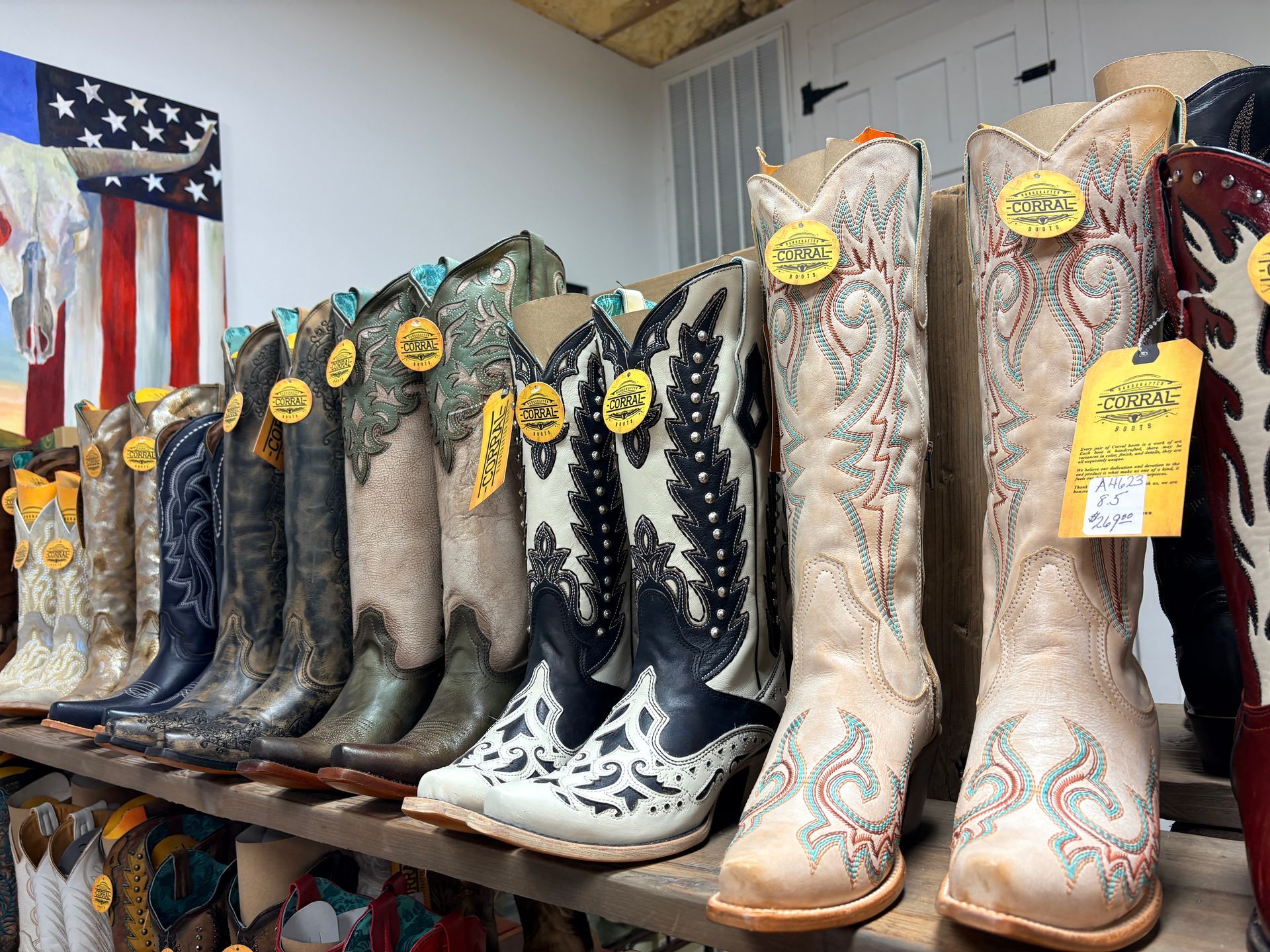 Row of decorated cowboy boots displayed on shelves, with yellow tags and an American flag in the background