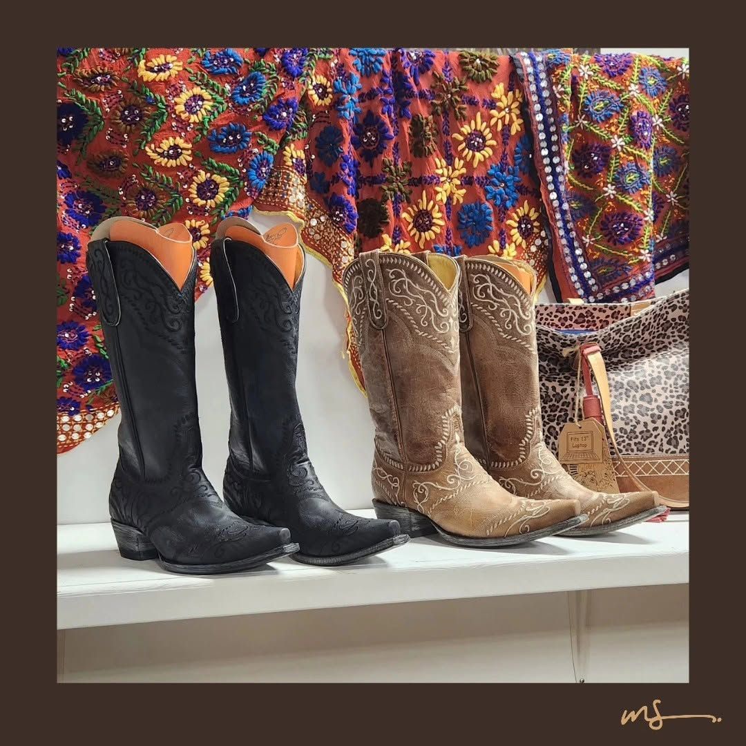 Two pairs of embroidered cowboy boots, black and tan, displayed on a shelf in front of a colorful textile backdrop