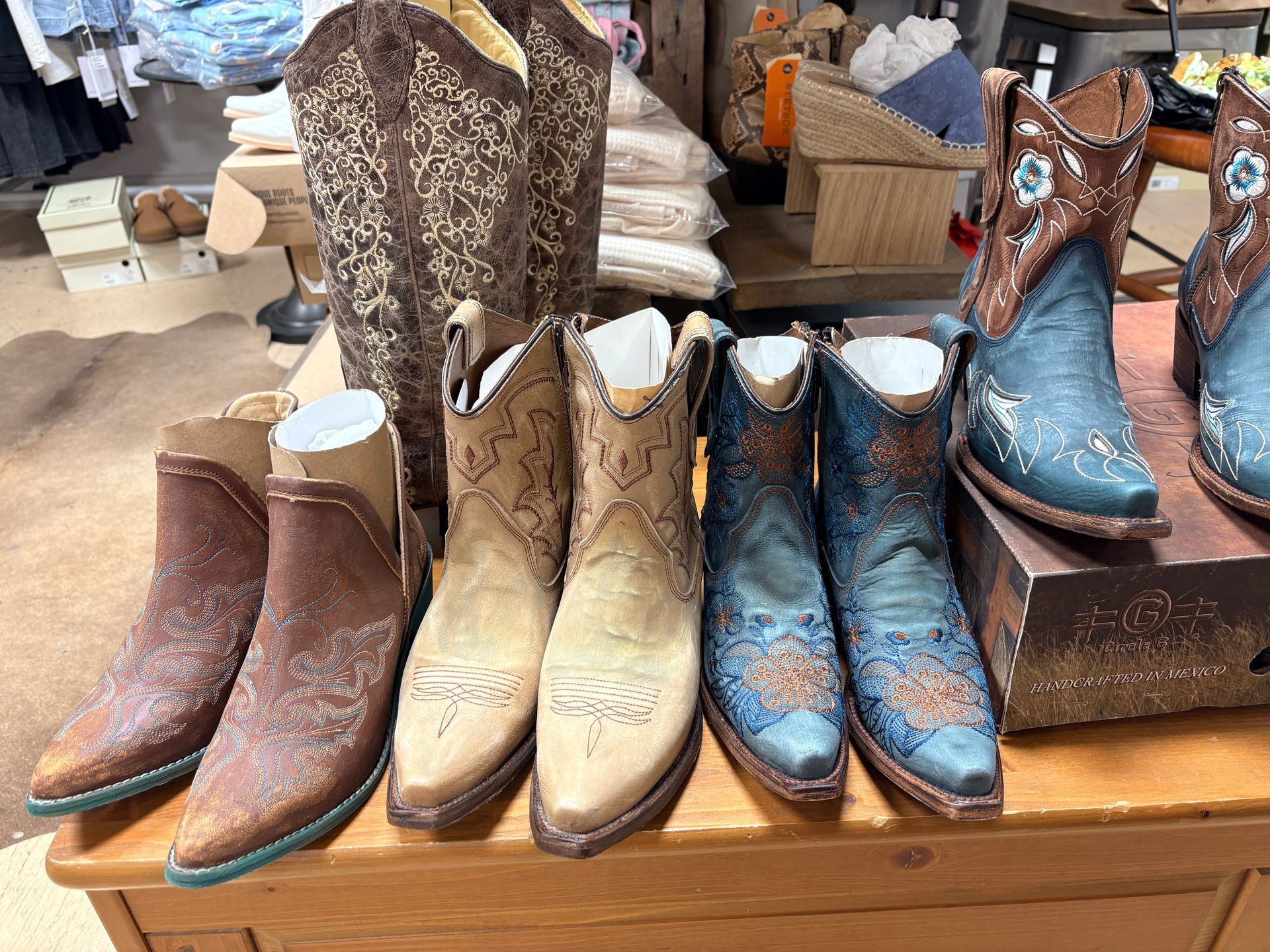 Display of cowboy boots in brown, beige, and blue on a store table, with more boots and shoes in the background
