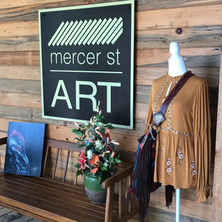 Mercer St ART sign beside a bench, floral arrangement, and tan fringed dress on a mannequin against wood wall