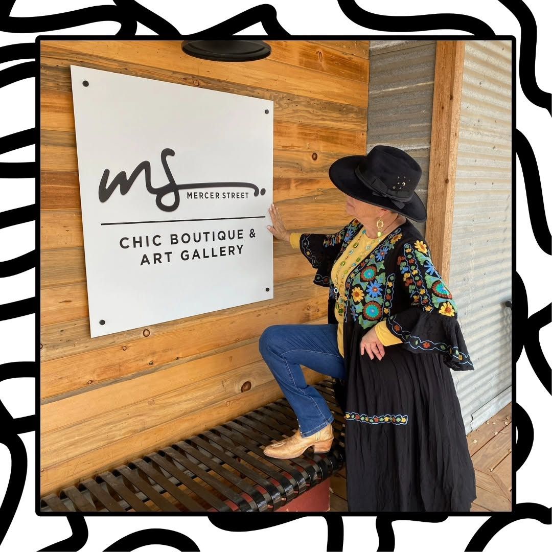 Woman in black hat posing by “Chic Boutique & Art Gallery” sign on a wooden wall.