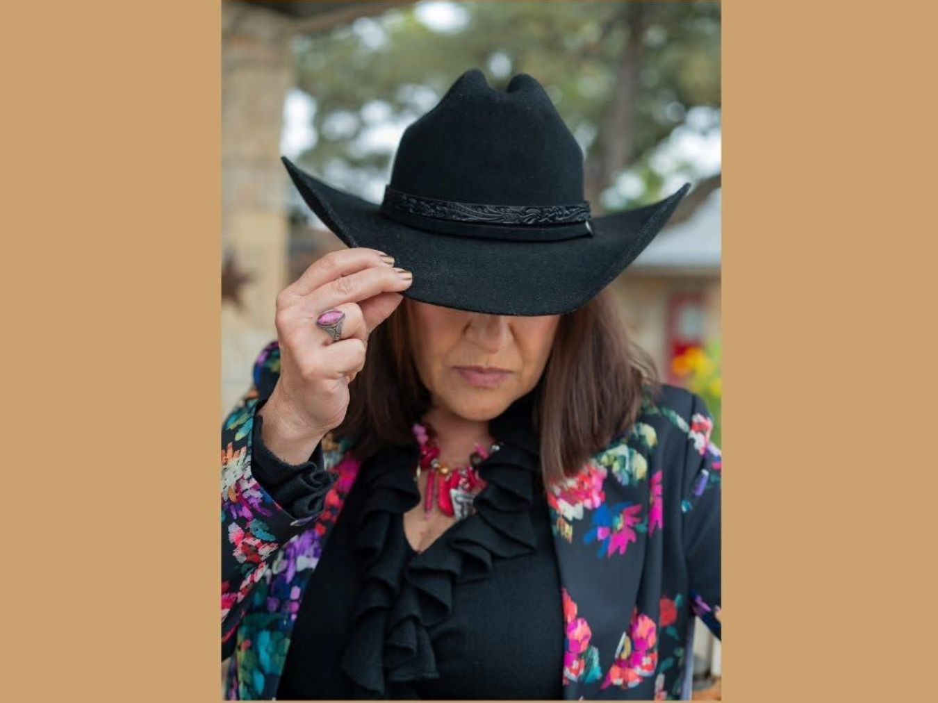 Woman in a black cowboy hat and floral jacket adjusting the brim outdoors