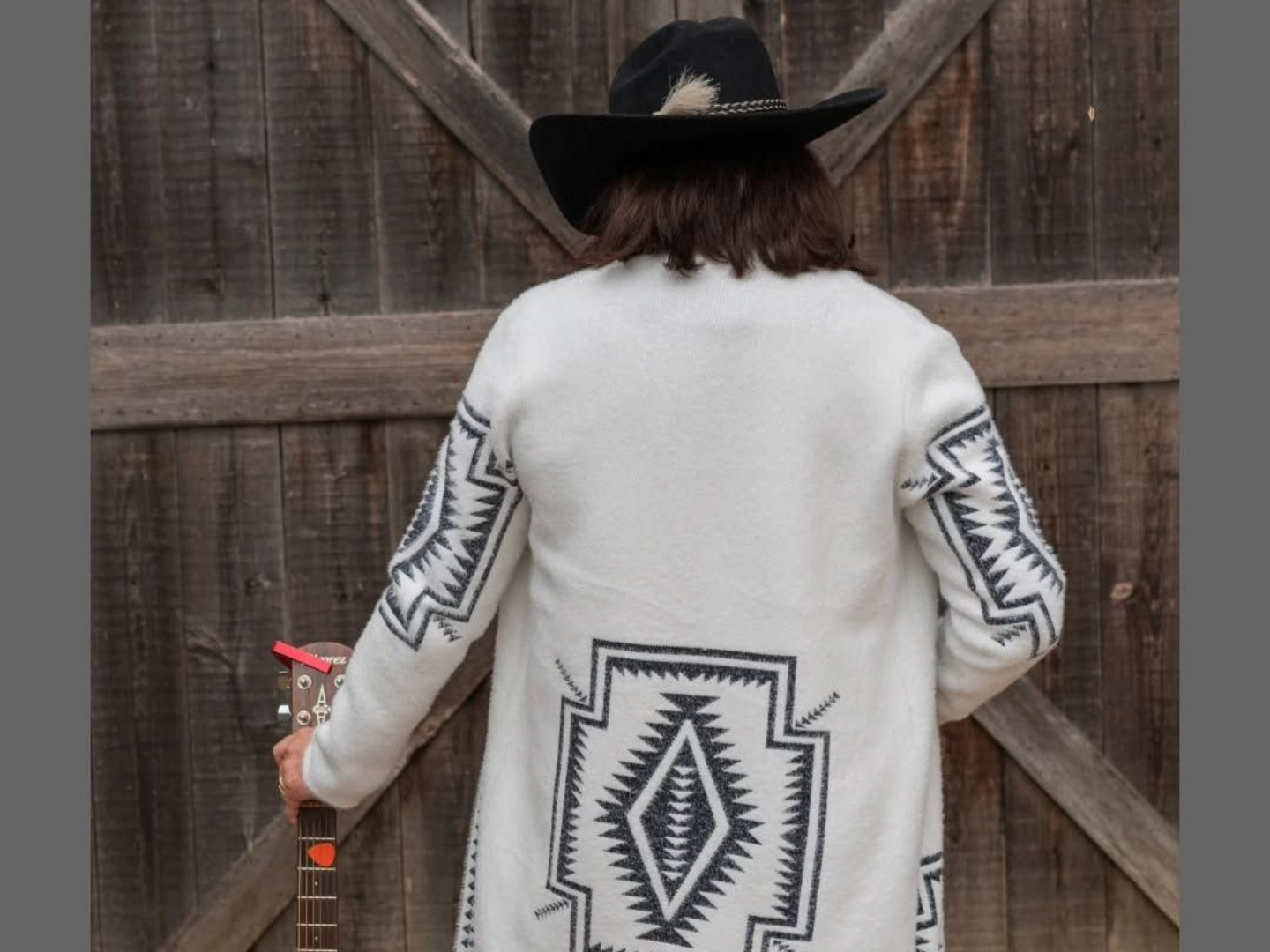 Person in a black hat and white patterned sweater facing a wooden gate, holding a red tool.