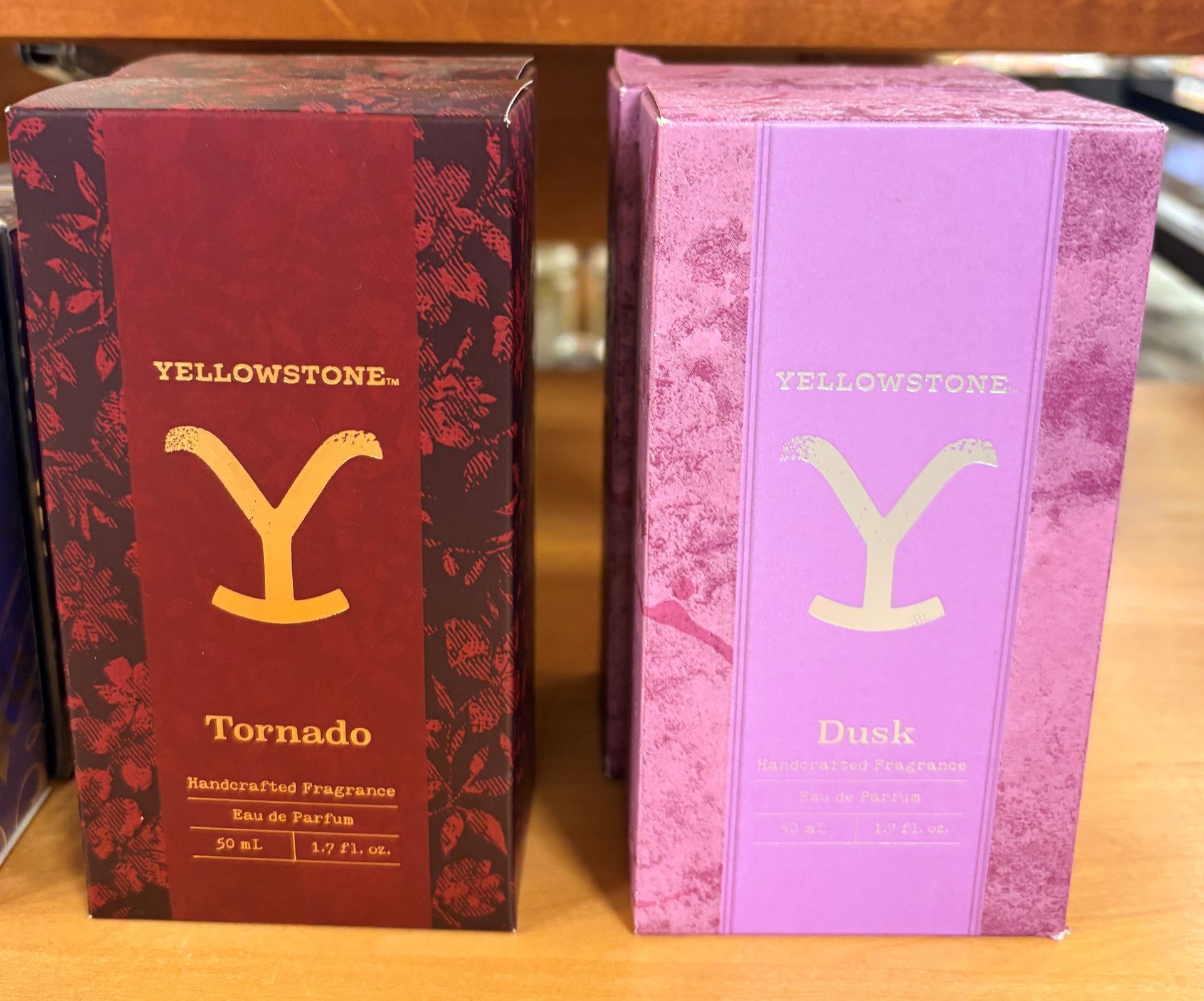 Two Yellowstone perfume boxes, one red “Tornado” and one pink “Dusk,” on a wooden shelf.
