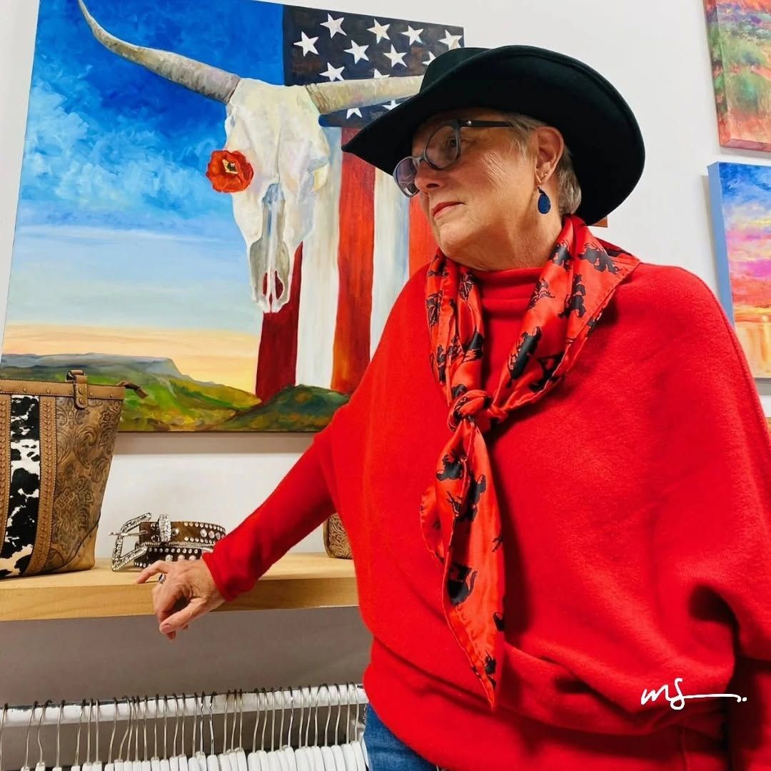 Person in a black hat and red scarf stands beside colorful artwork and a white skull display in a gallery.
