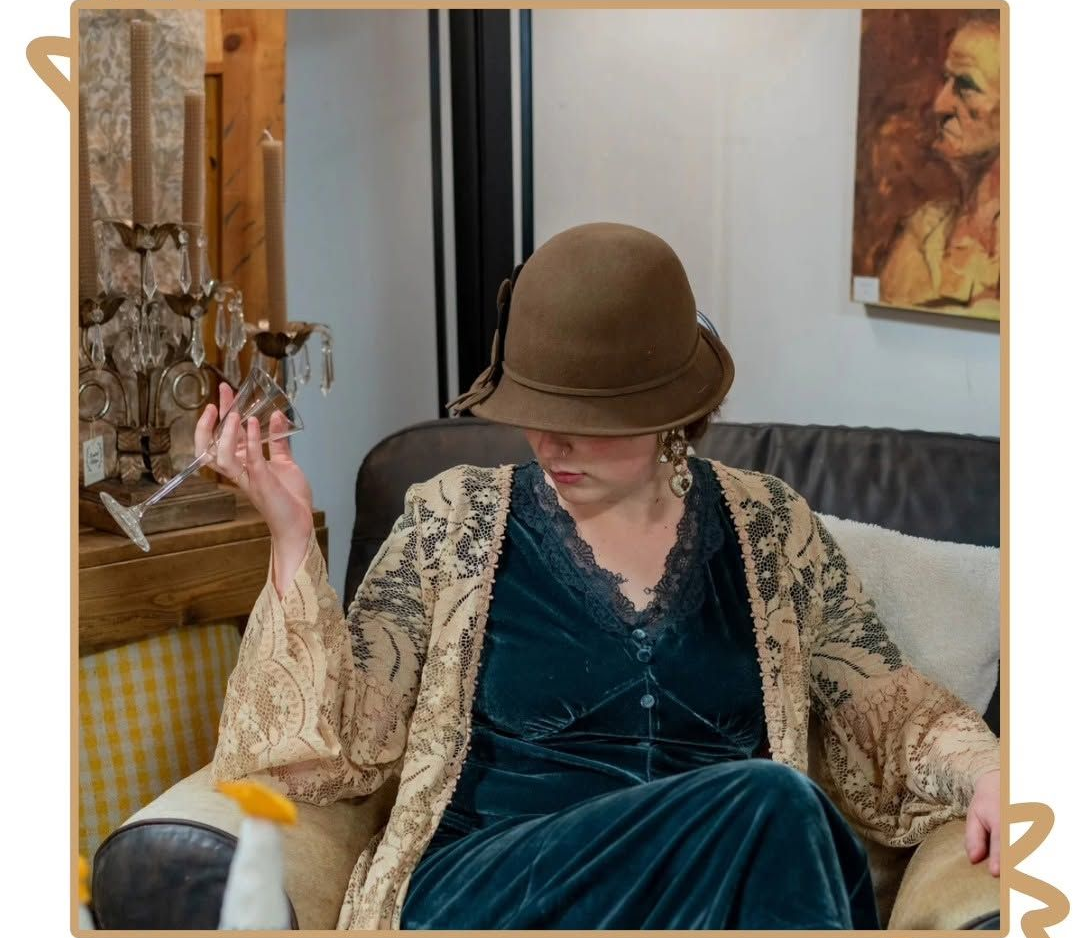 Person seated indoors in a brown hat and teal dress, arm raised beside a yellow chair and painting.