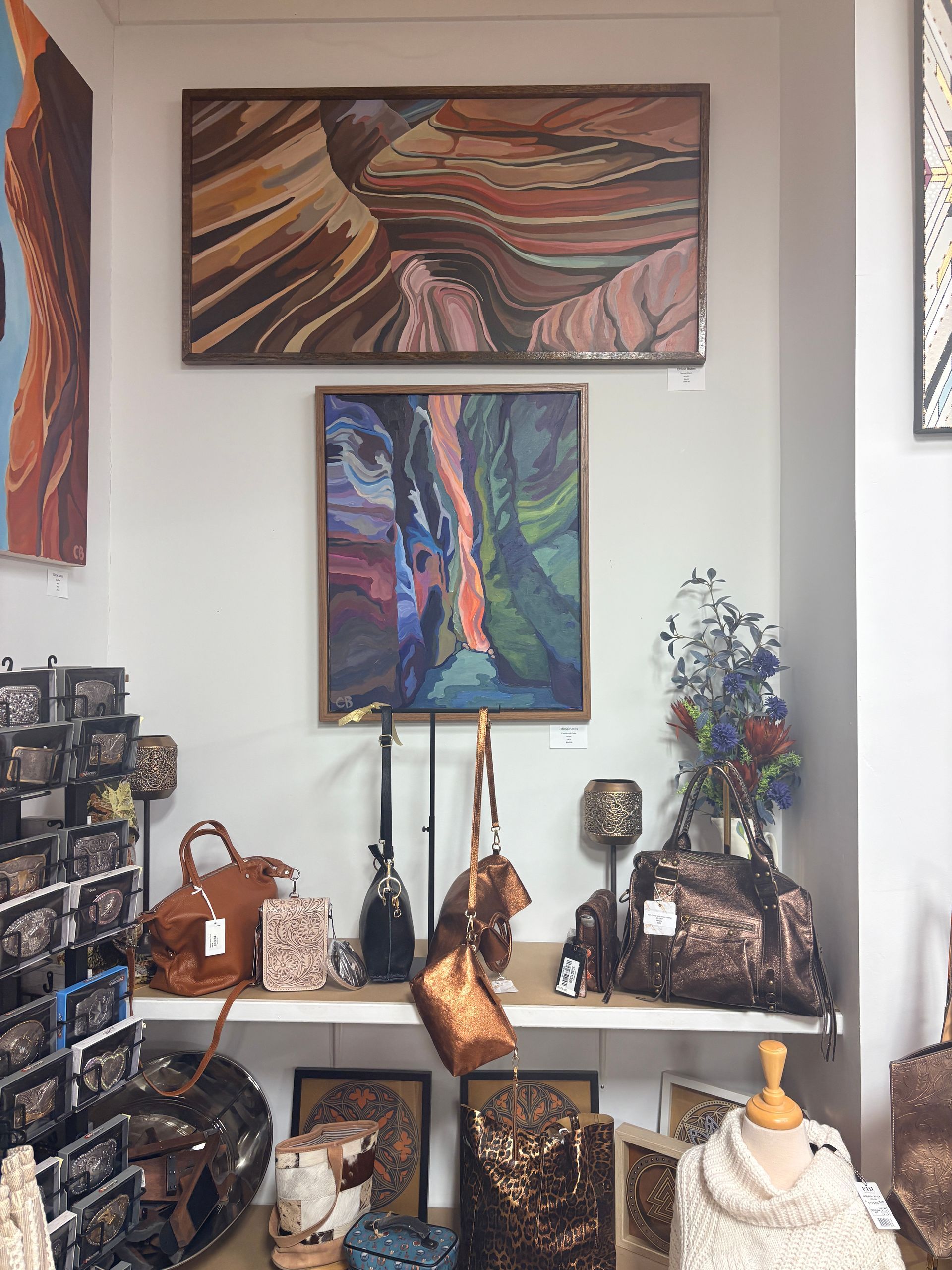 Art display of colorful paintings and handmade leather bags on shelves in a boutique-like room
