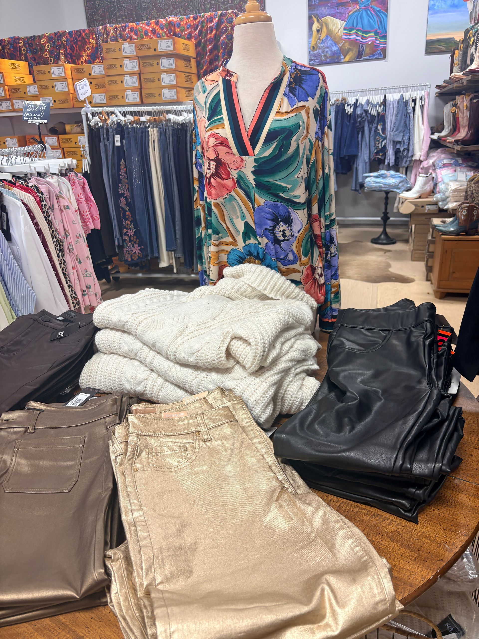 Clothing boutique display with folded sweaters and dresses on a round table, surrounded by hanging garments.