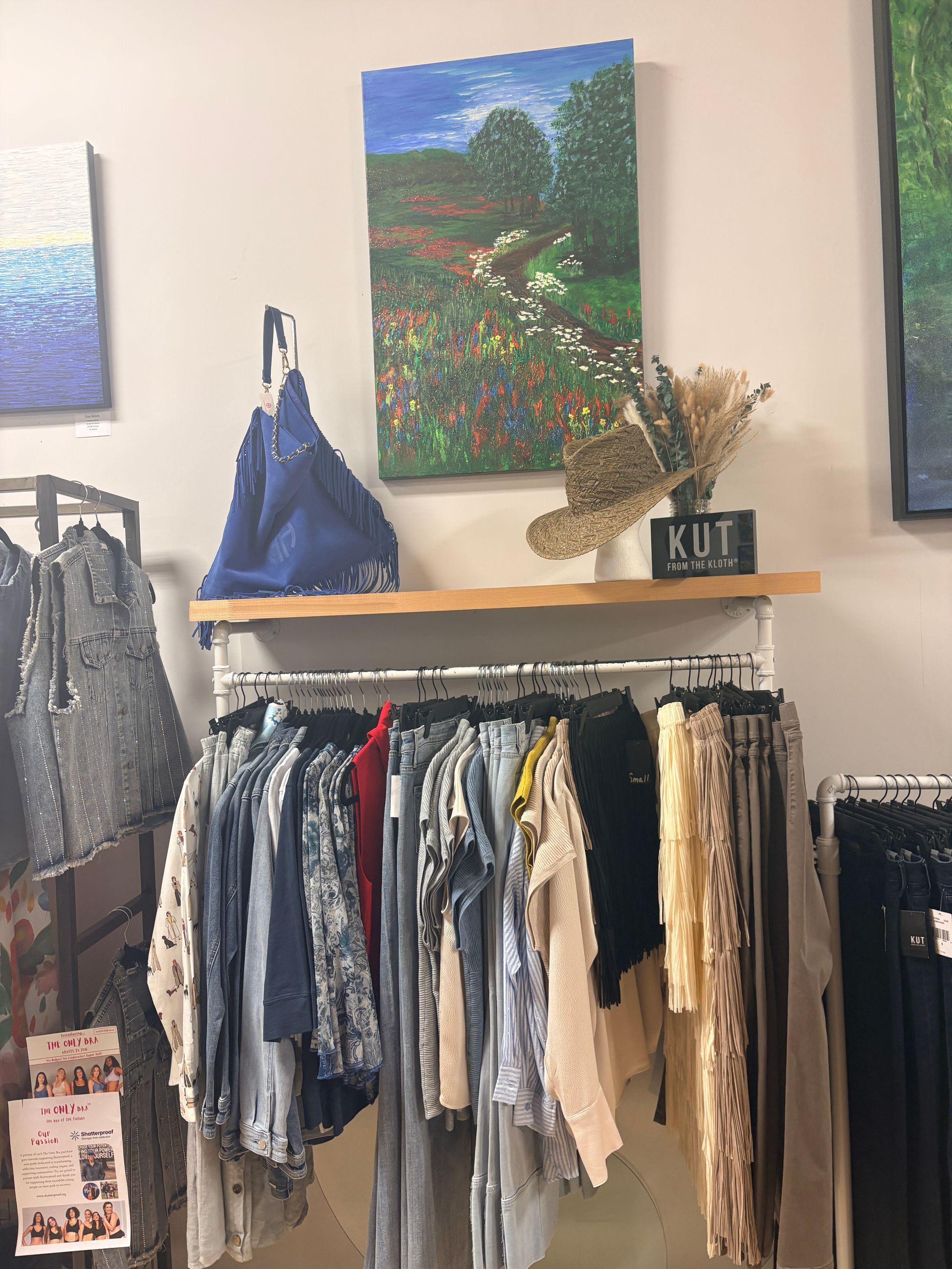 Boutique clothing rack with hanging tops, handbags, and wall art in a small shop interior