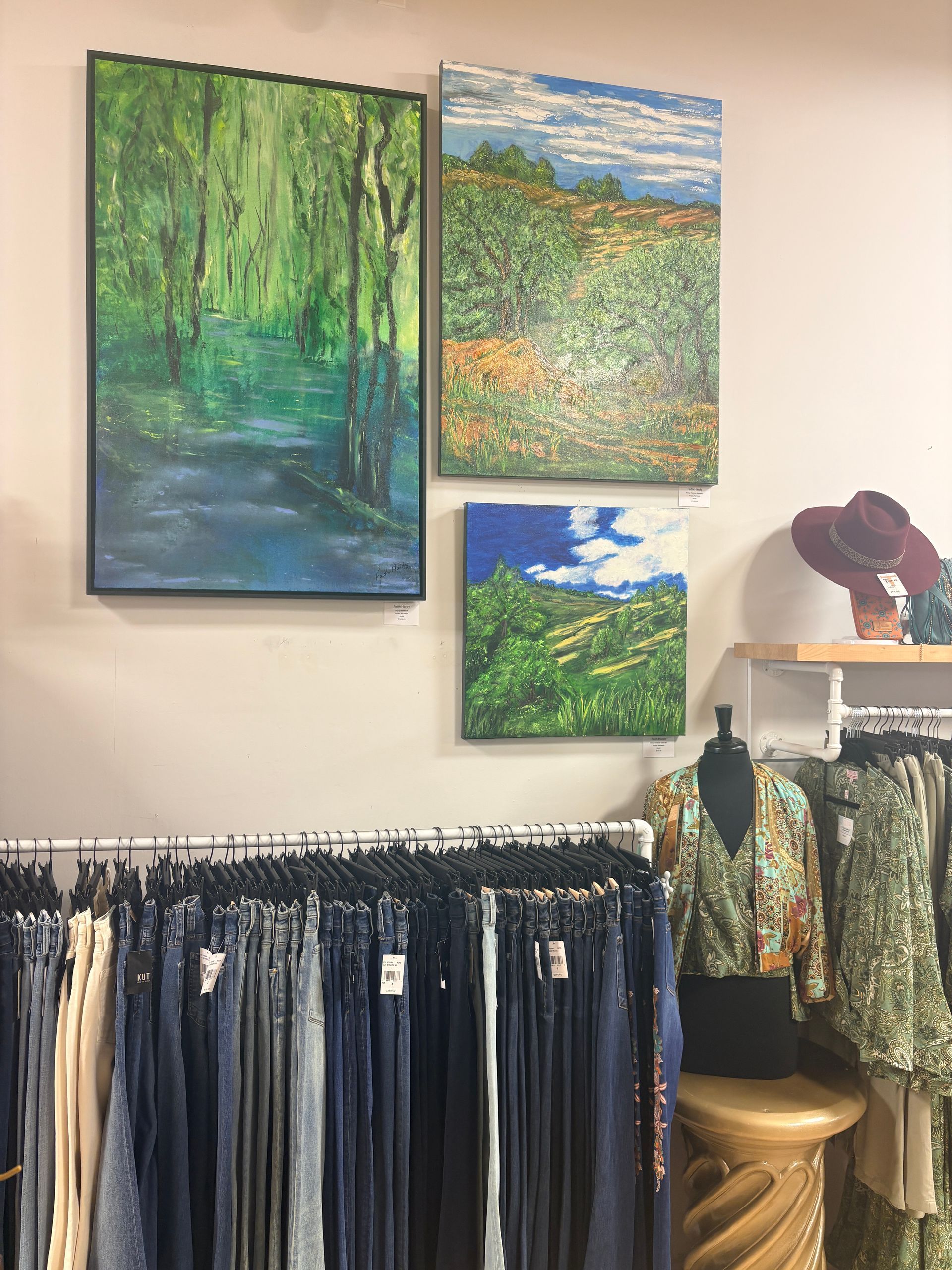 Art gallery display with landscape paintings above racks of denim and patterned clothing