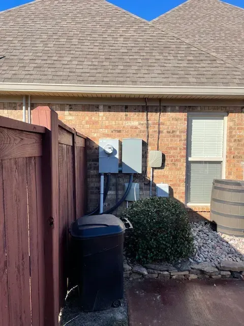 Electrical boxes and utility lines on a brick wall next to a fence and a trash can, under a brown roof.