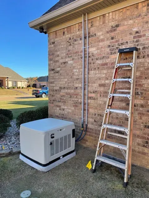 Generator next to a brick house. Ladder propped against wall, conduit pipe alongside. Clear sky.
