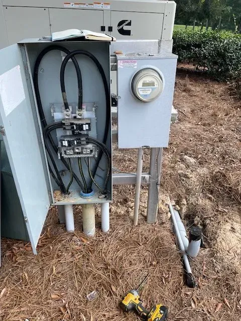 Electrical panel with exposed wiring, meter, and tools on ground. Exterior setting.