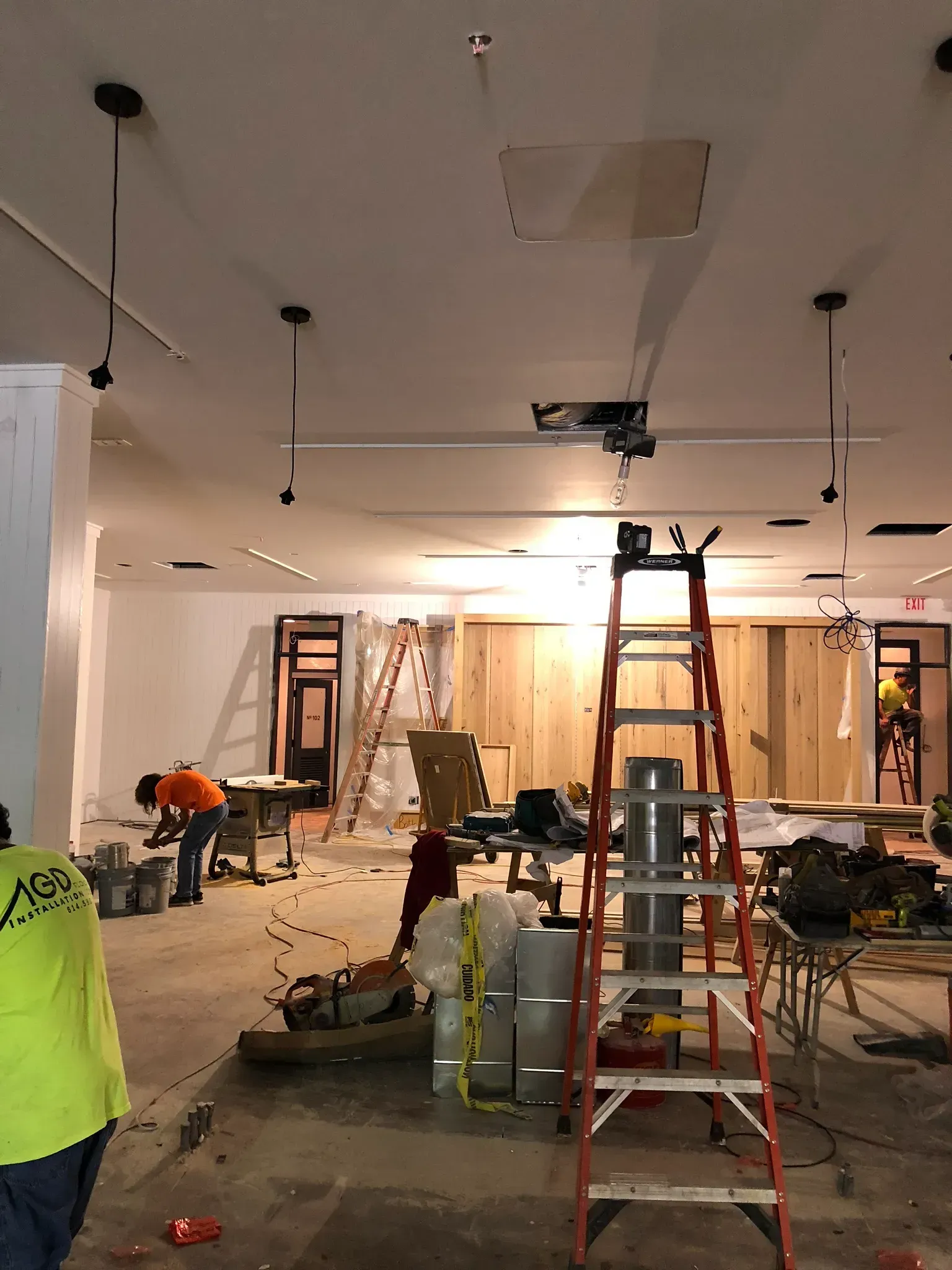 Construction in progress: workers, ladders, and tools in a room with wood paneling and lighting fixtures.