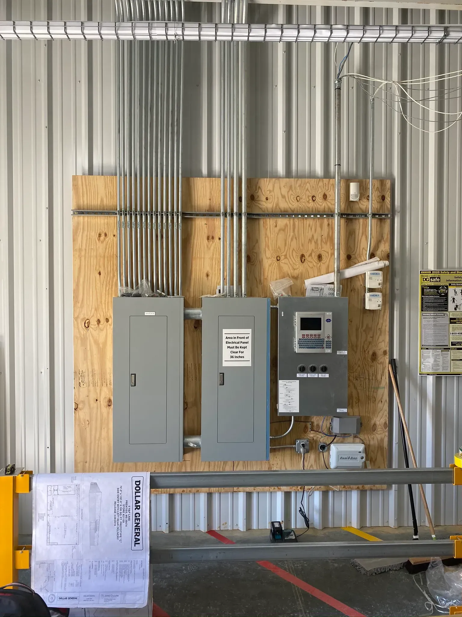 Electrical panels mounted on plywood in a metal building, with conduit and a schematic in front.