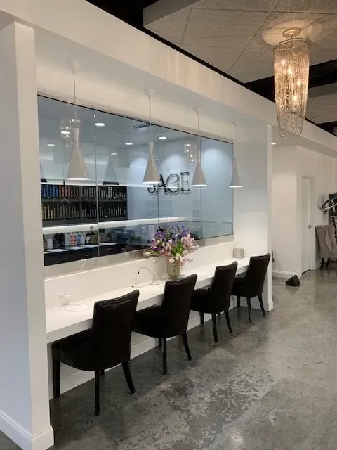 A row of black chairs at a white counter with a glass window behind, inside a well-lit salon.