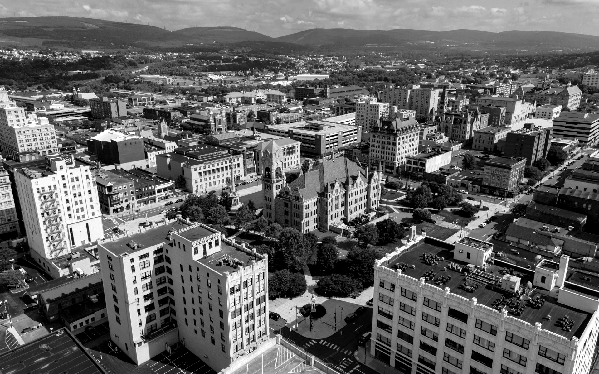 Black and white aerial view of a city with tall buildings, mountains in the background, and a bright sky.