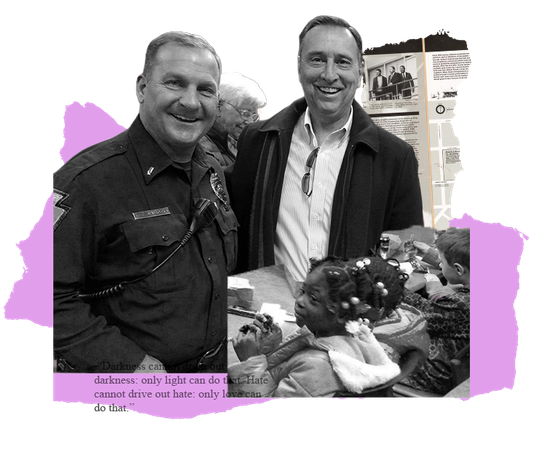 Two men smiling, one in a uniform.  They stand near a table with objects.