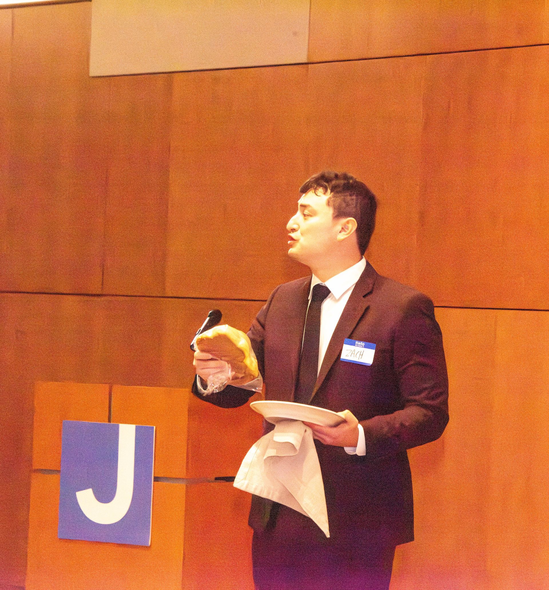 Man in a suit speaking at a podium, holding a bread roll and plate. Room with orange wall and 
