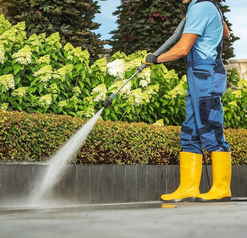 Pressure Washing A man in blue overalls and yellow boots is using a high pressure washer to clean a sidewalk.