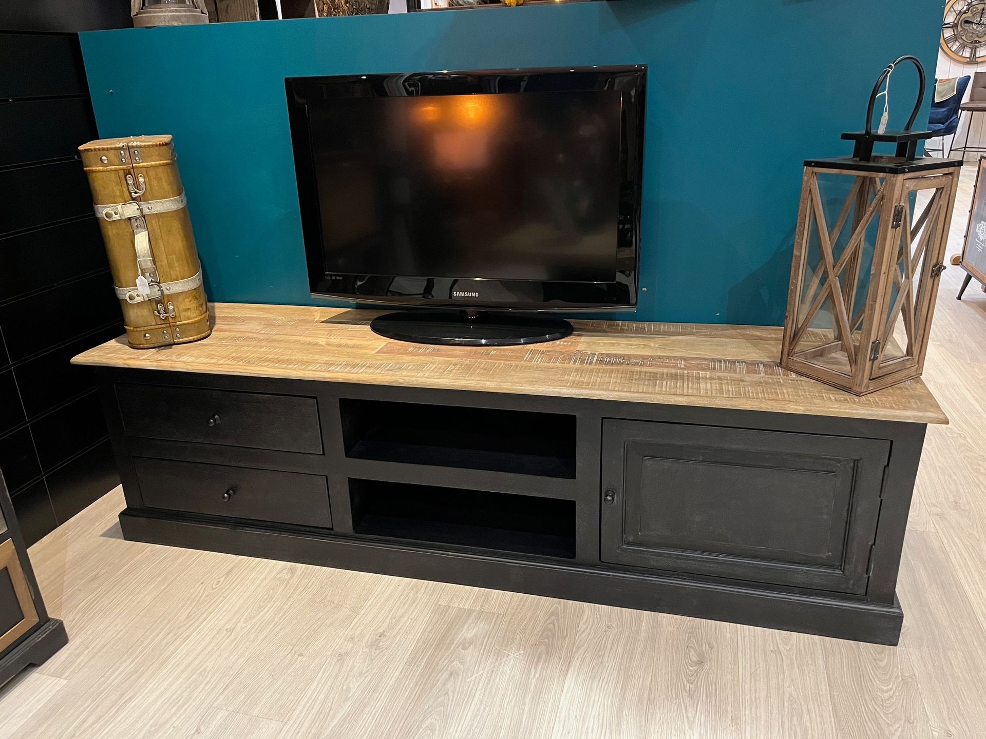 Meuble TV noir avec un téléviseur dessus, mur bleu turquoise en arrière-plan. Décoration : une lanterne et un bocal en métal.
