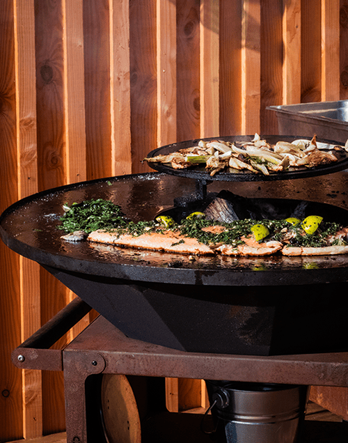 Poste de cuisson extérieur avec un bol de cuisson noir, une étagère de rangement en bois et un fond en bois.