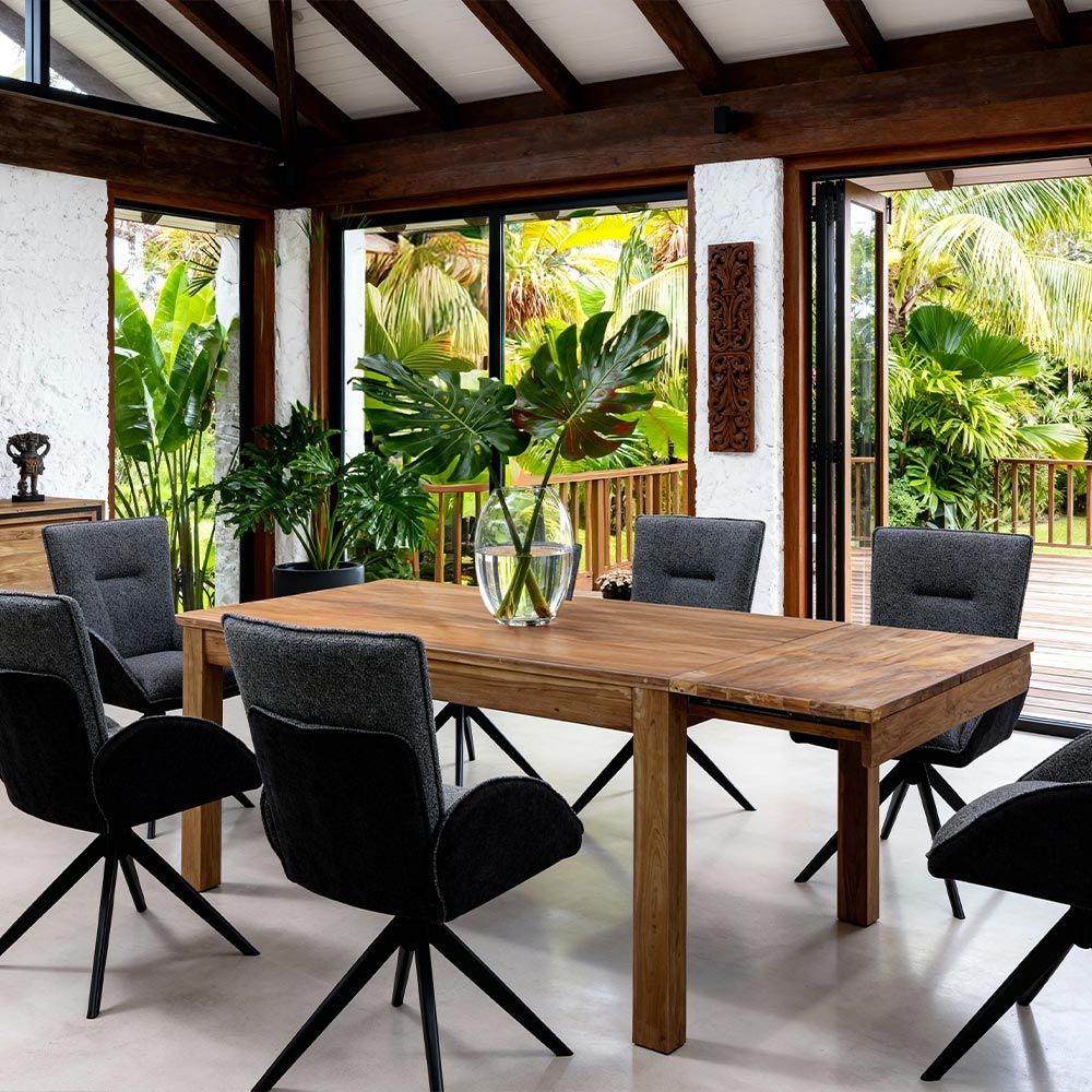 Salle à manger moderne avec table en bois, chaises noires et grandes fenêtres donnant sur une végétation tropicale luxuriante.