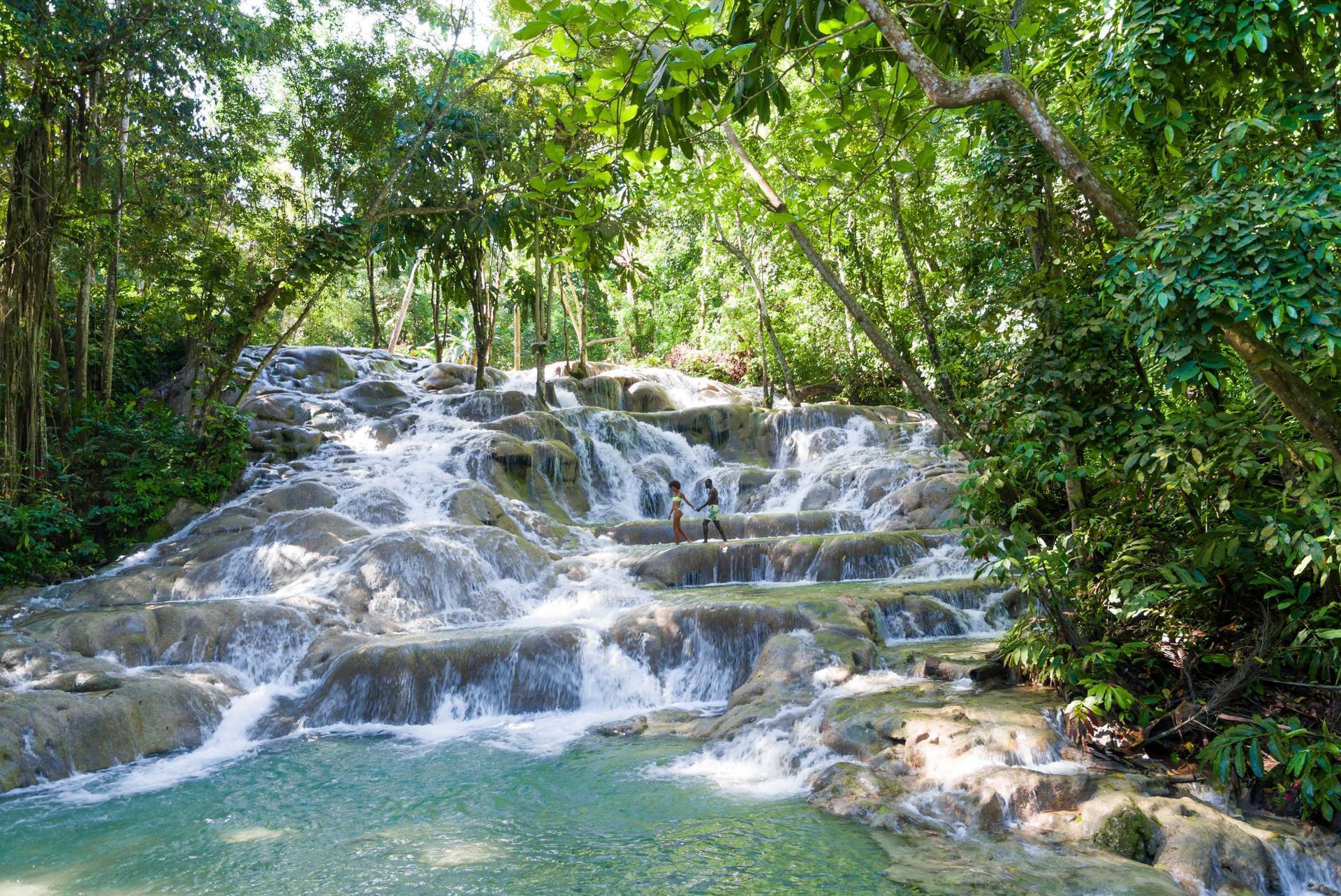 Tiered waterfall flowing through lush green forest, with turquoise water and white cascades over rocks.