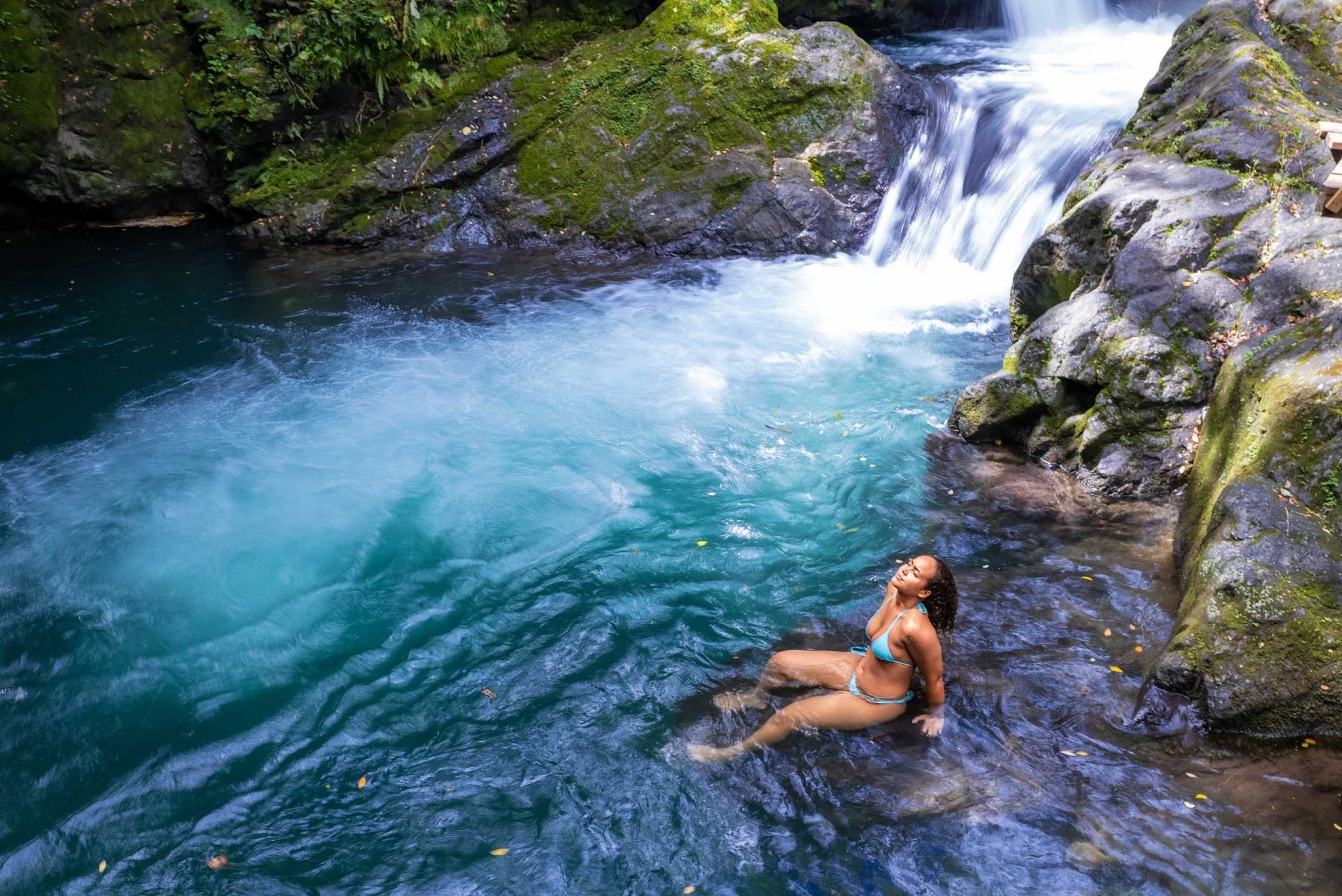 Person relaxing in a clear blue river pool beside a small waterfall and mossy rocks