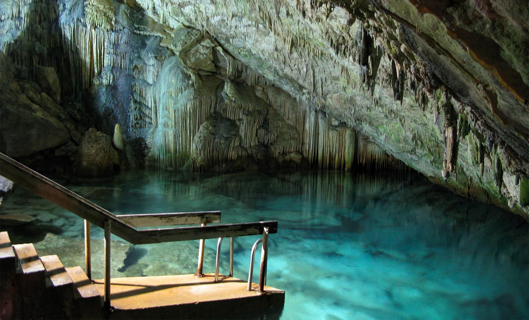 Turquoise cave pool with a wooden deck and staircase along a rocky, sunlit cavern wall 500K year old Green Grotto Caves
