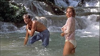 Two people wading in a rushing river near a rocky waterfall, looking back toward the camera.