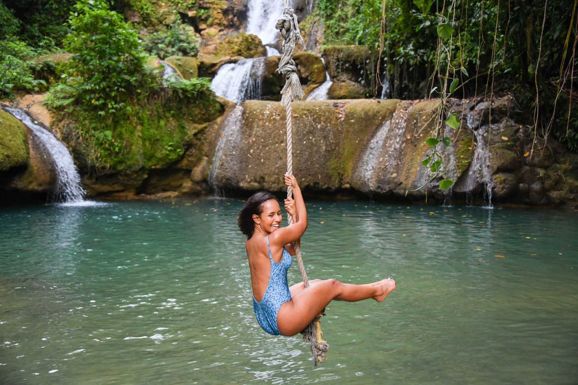 Person in blue swimsuit swinging on a rope over a clear pool below a small waterfall
