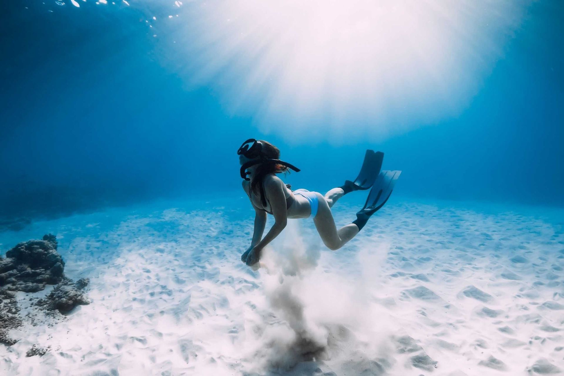 Scuba diver kneels on sandy seafloor with a ray under bright blue water and sun rays above