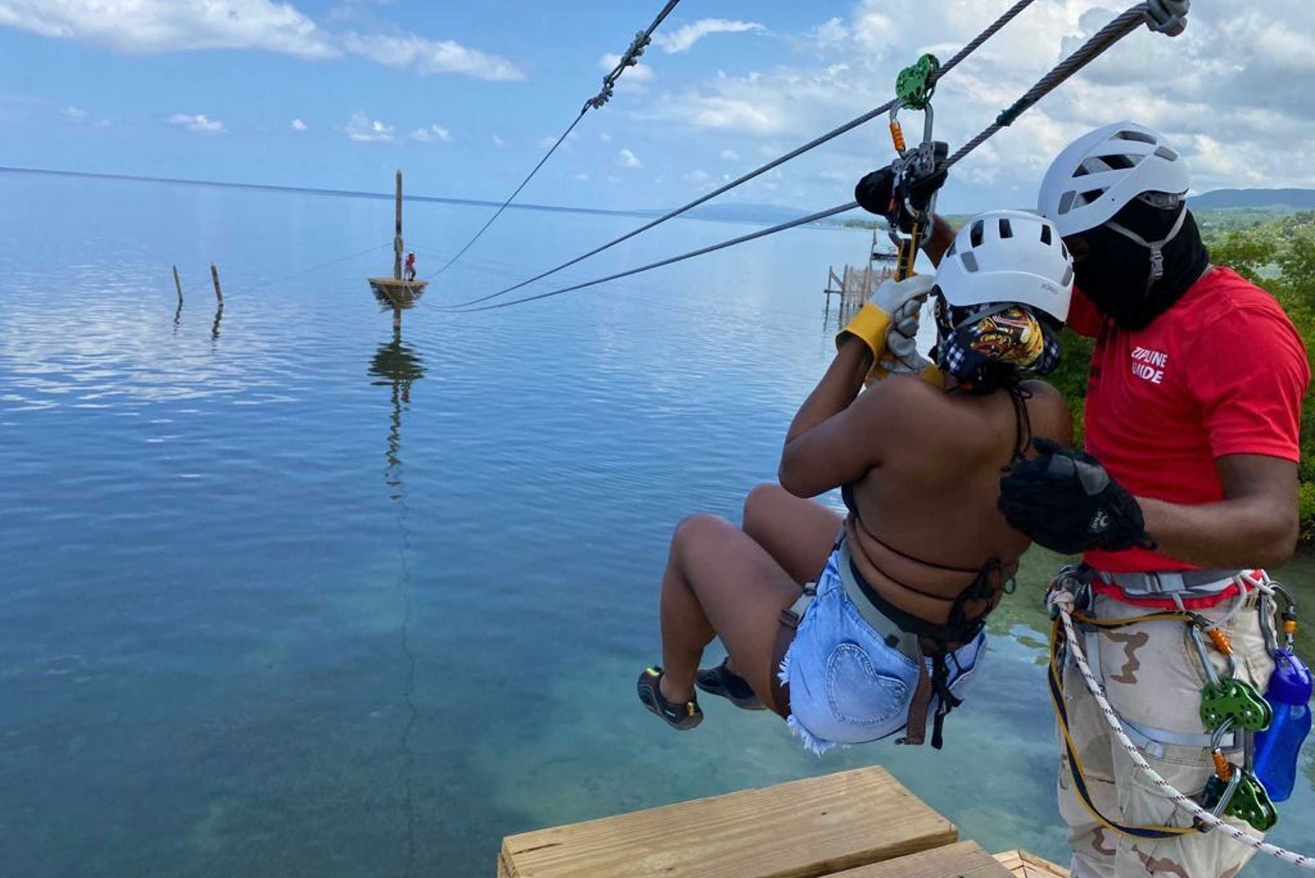 Harnessed zipline rider over calm blue water, guided by an instructor at a dock.