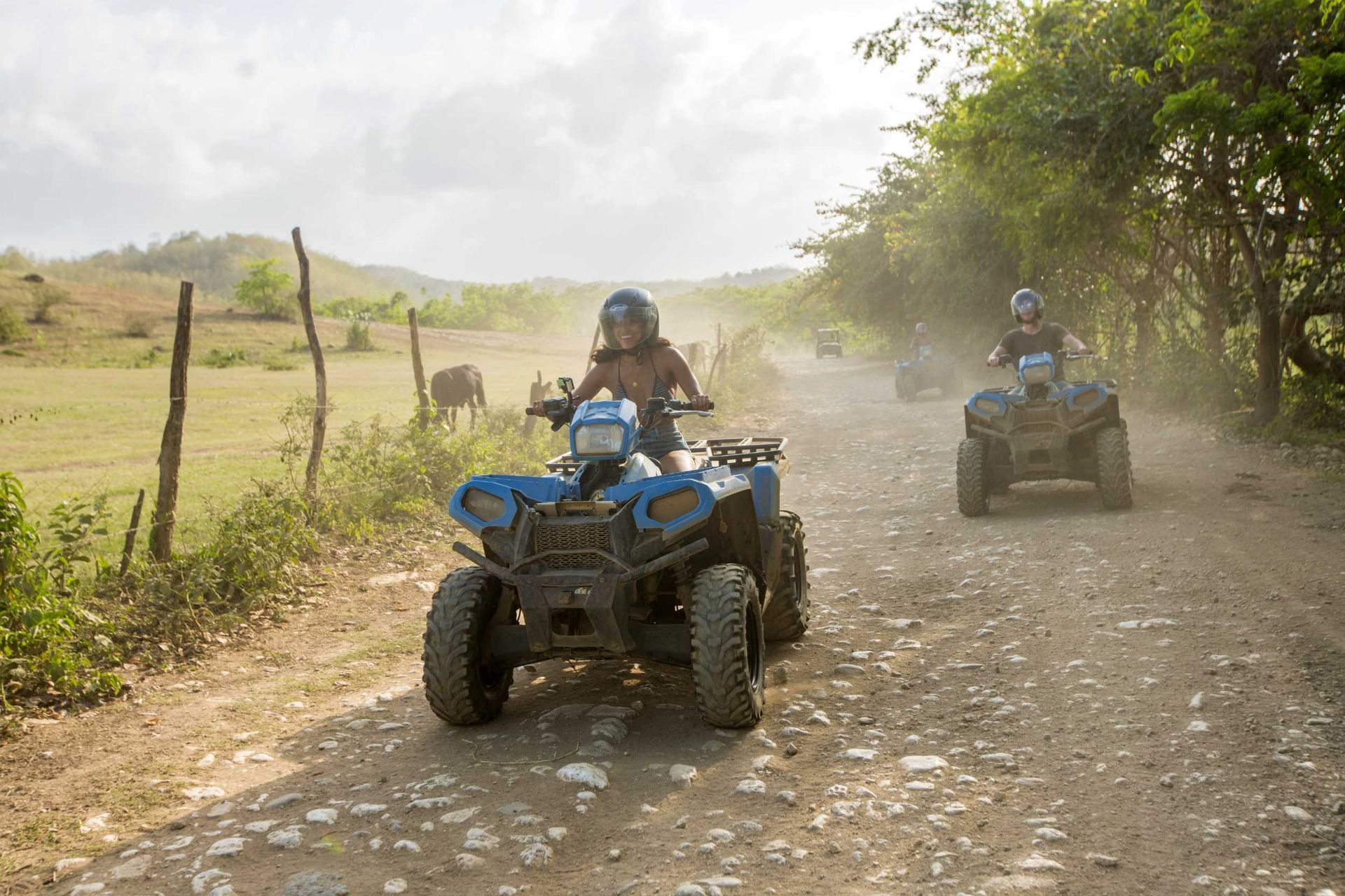 Two people ride blue ATVs on a dusty dirt road in a rural field.